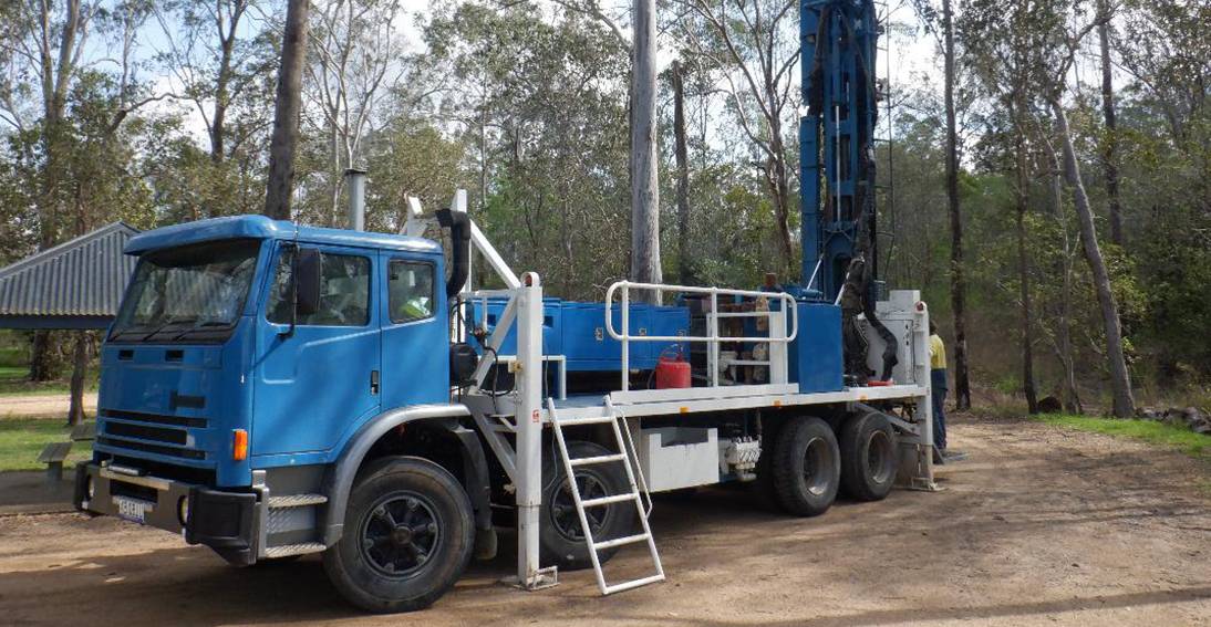 A Blue Drilling Truck Equipped with A Large Vertical Drill Parked in A Rural Area with Trees and A Metal-Roofed Building in The Background — Bore Drilling in Bundaberg, QLD
